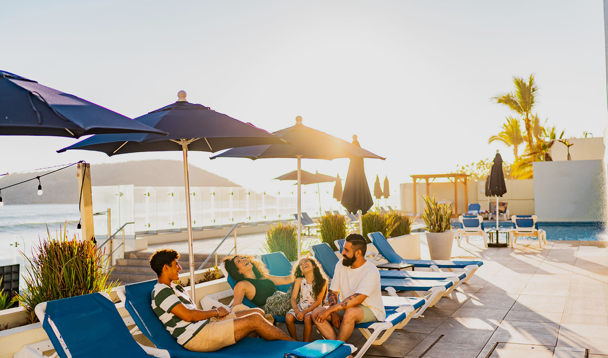 Family relaxing by pool under blue umbrellas.